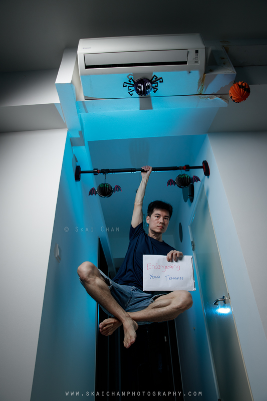 Skai hanging one-handed from a pull-up bar beneath the cooling unit while holding his handwritten Tengah protest sign